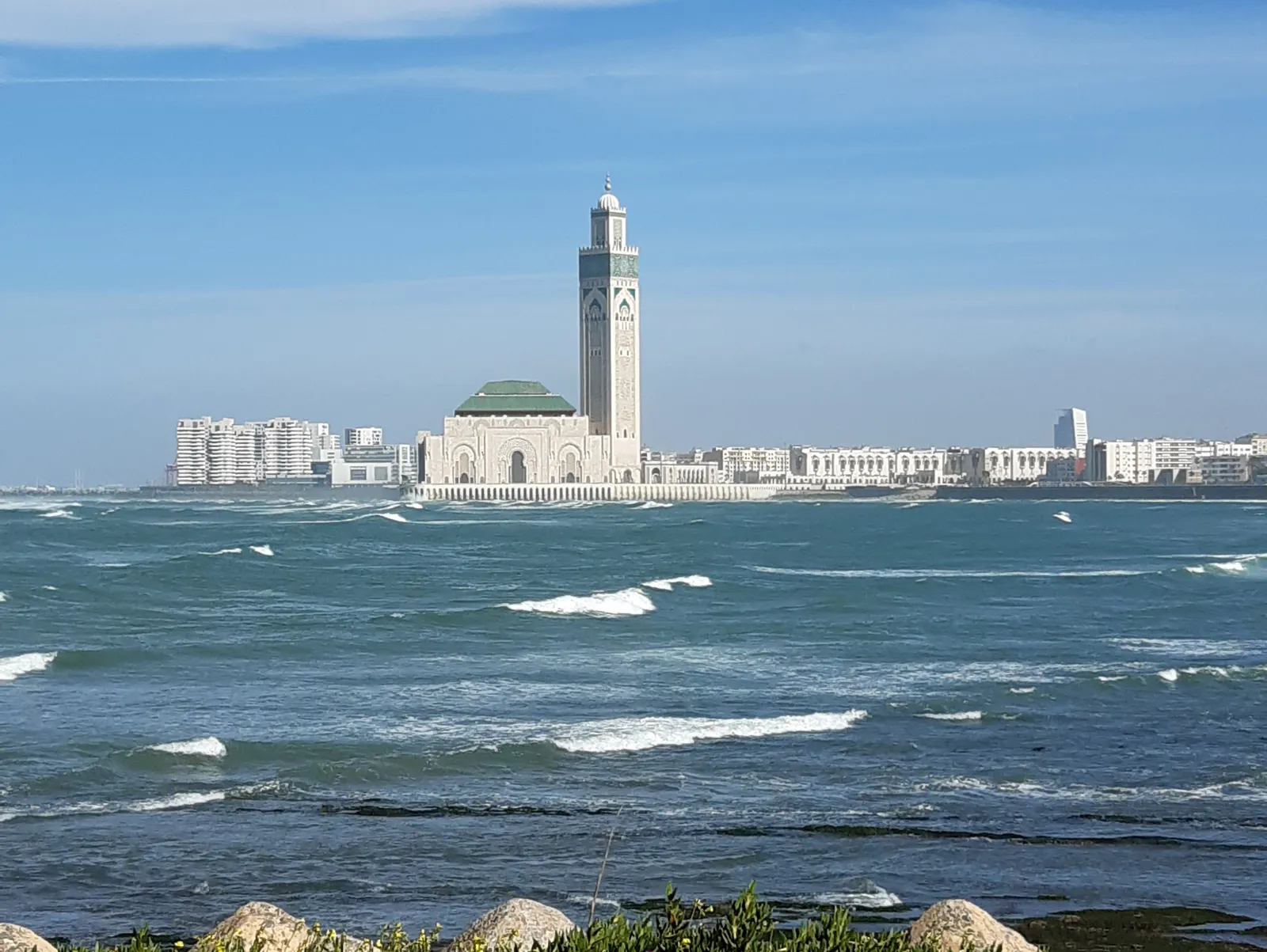 The Hassan II Mosque seen across the Atlantic, its green pyramidal roof and tall square minaret rising from an arcaded base at the water's edge, with the white apartment blocks of Casablanca along the shore behind