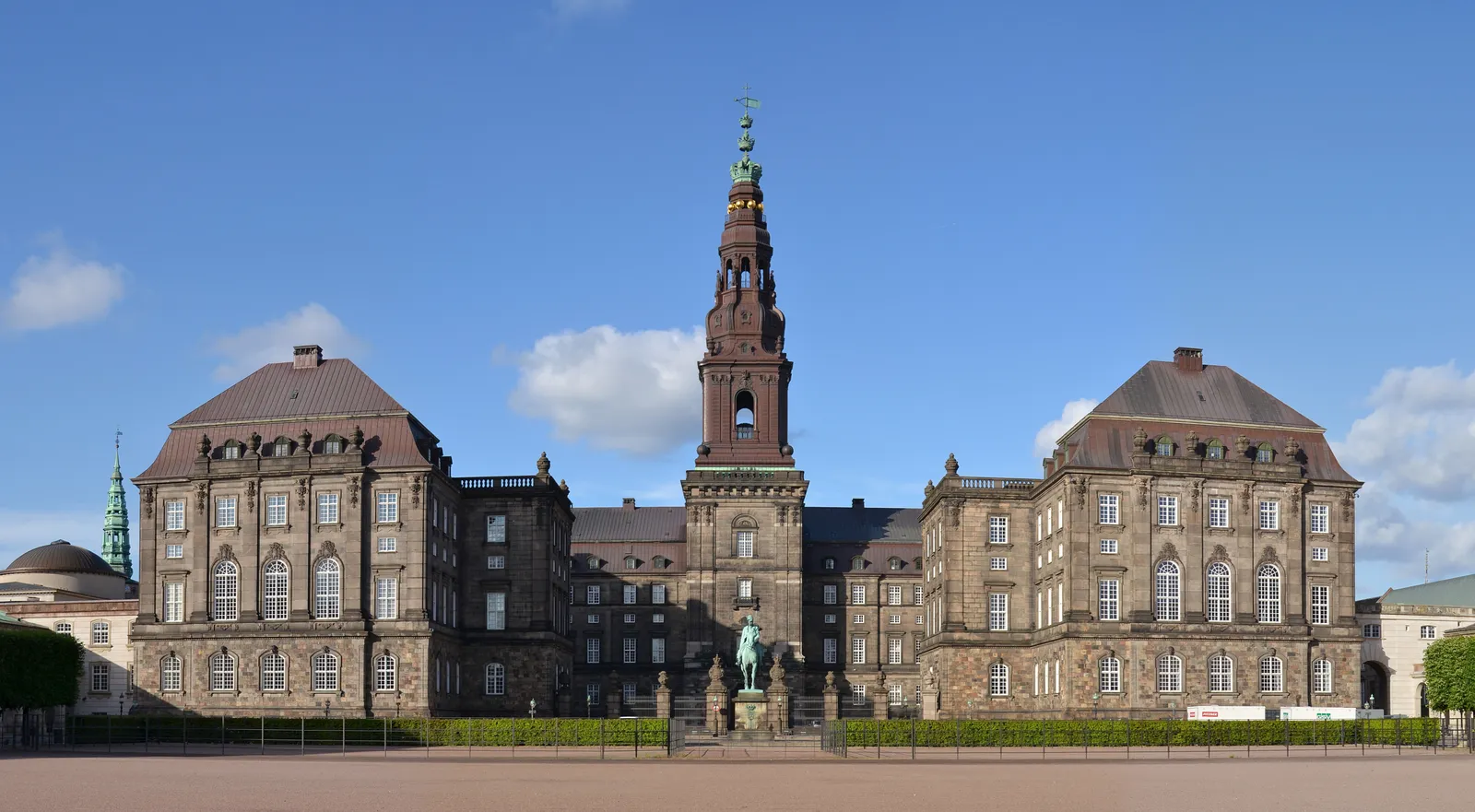 The front of Christiansborg Palace in Copenhagen seen across its cobbled square, two long grey wings either side of a central block, with the tall brick tower and its green copper spire rising over the middle