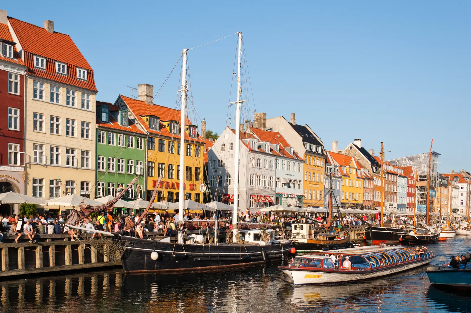The north side of Nyhavn in Copenhagen, a row of narrow townhouses painted red, yellow, blue and white above quayside awnings, with wooden sailing ships and a low tour boat moored in the canal in front of them