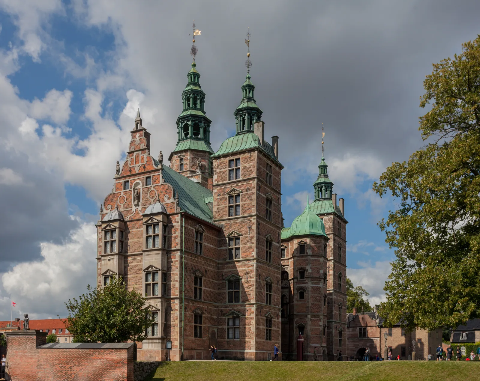Rosenborg Castle in Copenhagen, a red brick Renaissance house with sandstone banding and three green copper spires, standing behind a low brick wall and a strip of lawn under a cloudy sky