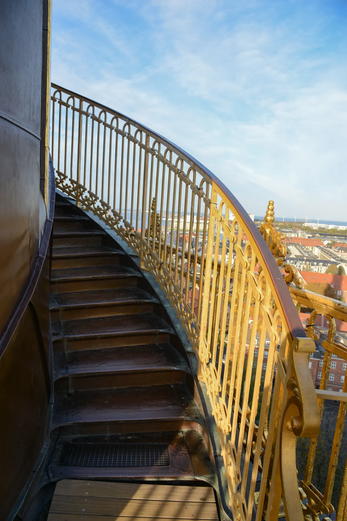 The outdoor staircase on the spire of the Church of Our Saviour in Copenhagen, dark treads curving around the outside of the spire behind a gilded handrail, with the rooftops of the city and the water far below