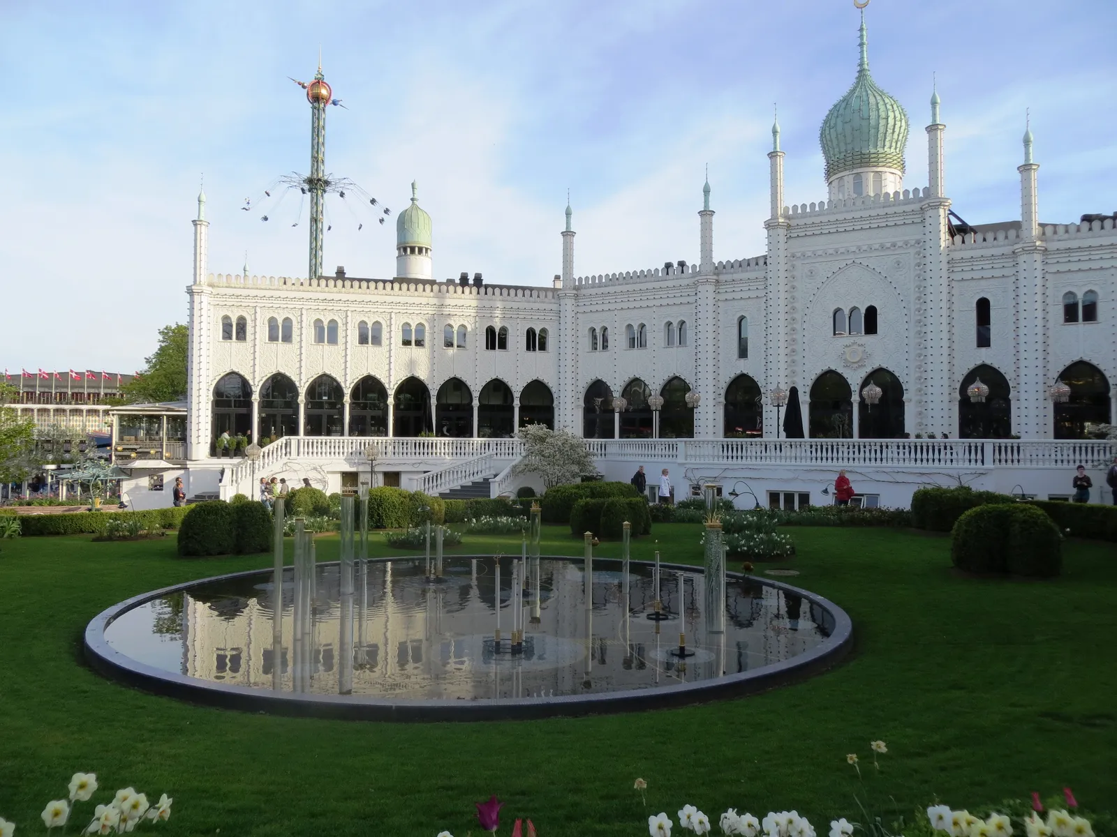 The white Moorish-style Nimb building in Tivoli Gardens, Copenhagen, its green onion dome and slim minarets above an arcaded front, with a lawn, flower beds and a low fountain pool in front of it