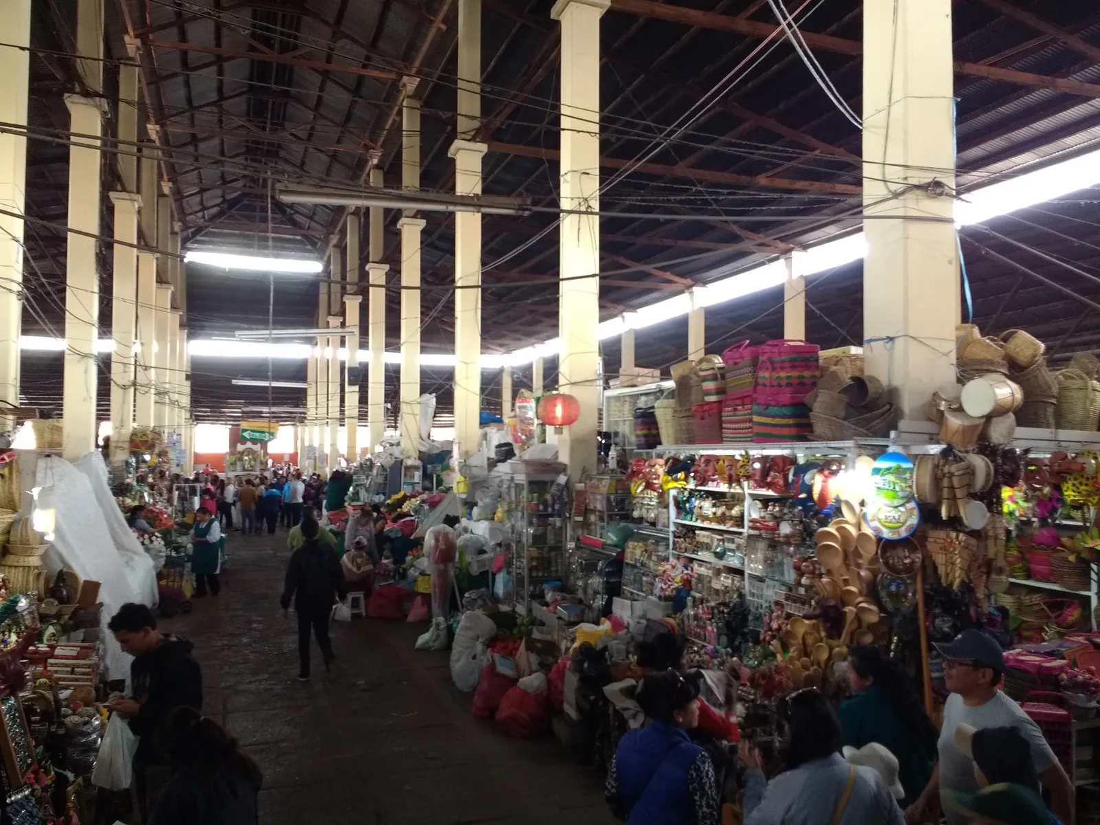 Inside San Pedro market in Cusco, a wide aisle running between craft and produce stalls under a high corrugated roof carried on rows of square columns