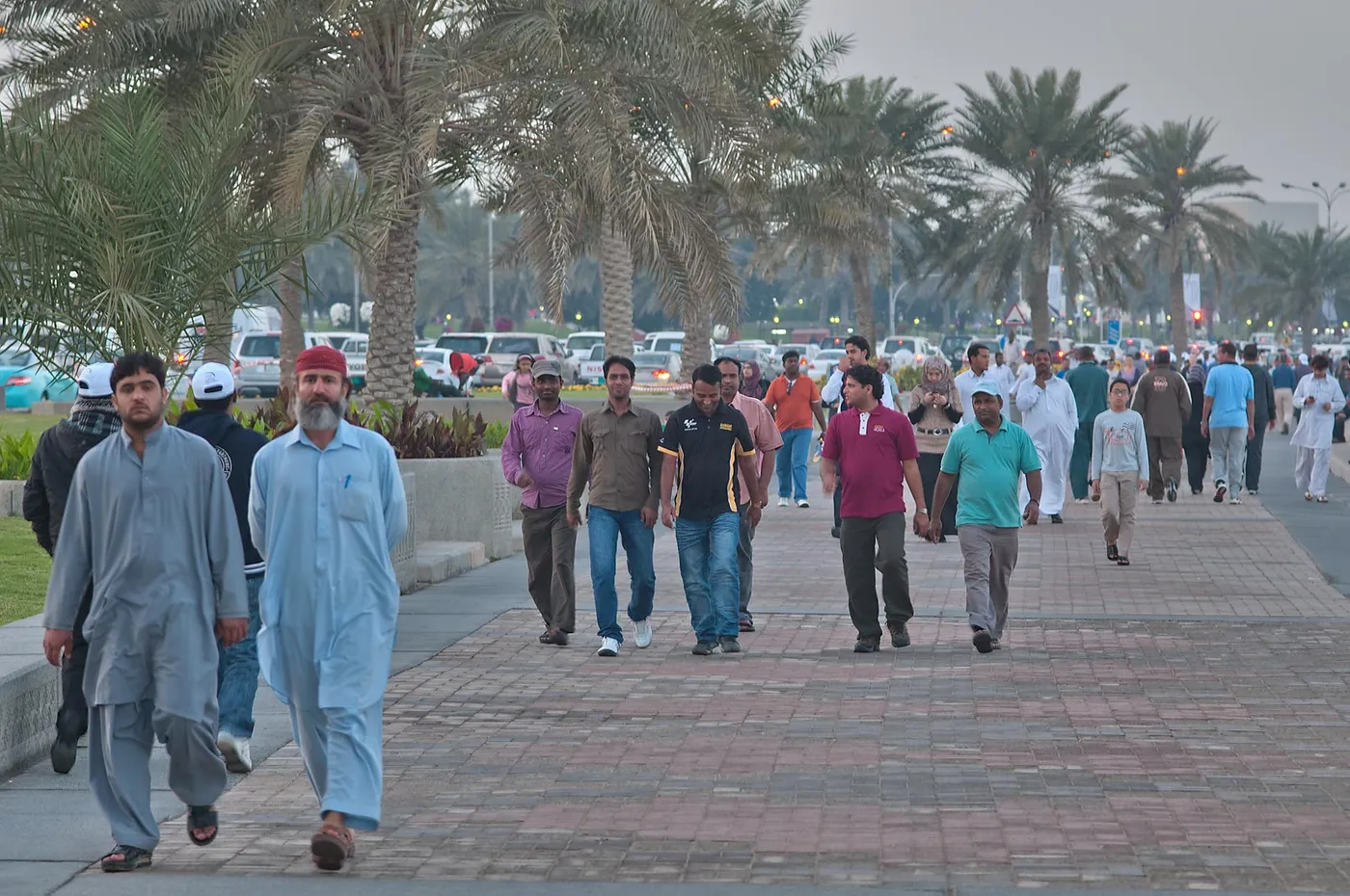 People walking along the brick-paved promenade of the Doha Corniche under a line of palm trees