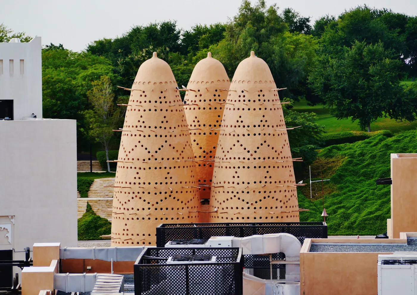Three tall conical pigeon towers at Katara Cultural Village in Doha, built of pale clay and pierced with rows of nesting holes, with a green planted bank behind them