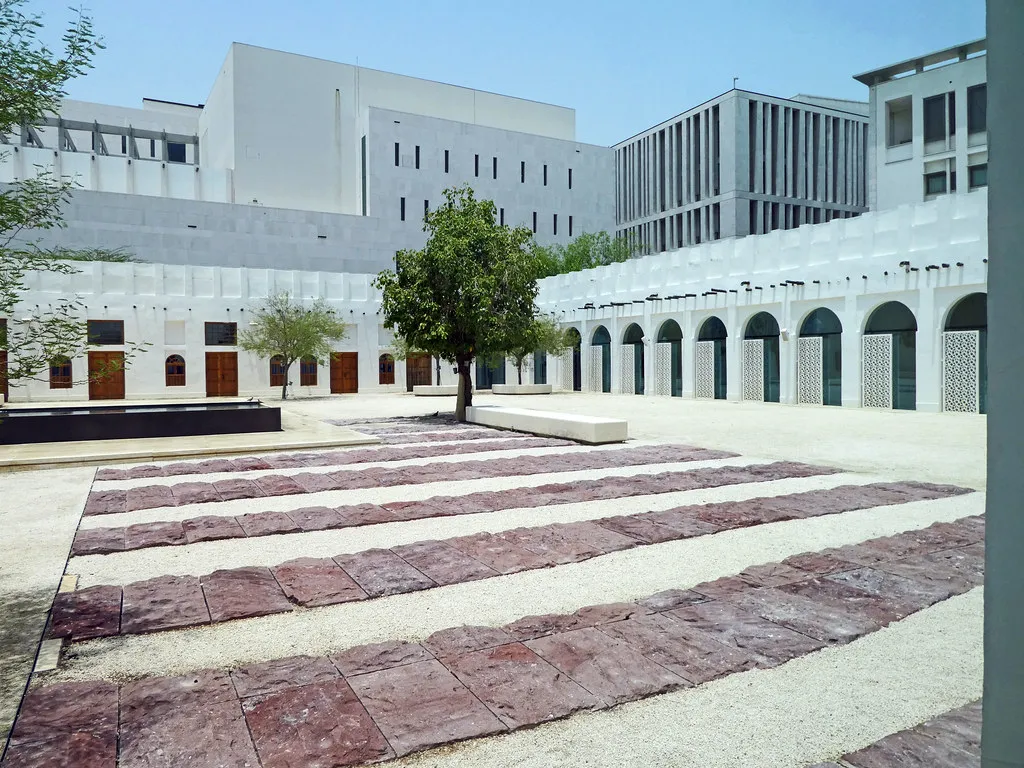 The white arcaded courtyard of the Msheireb Museums in Doha, a single tree in the middle of a paved yard with the blocks of Msheireb Downtown rising behind