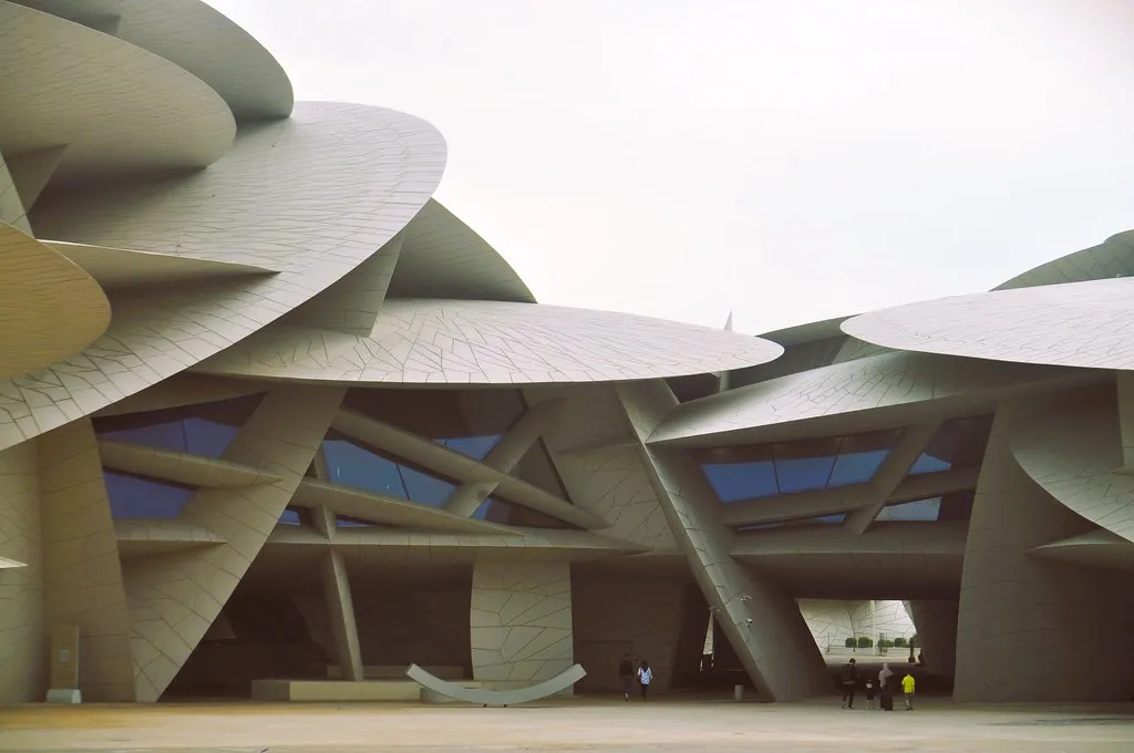 The National Museum of Qatar seen from its forecourt, huge sand-coloured discs tilted and interlocking above a glazed ground floor, with visitors small beneath them