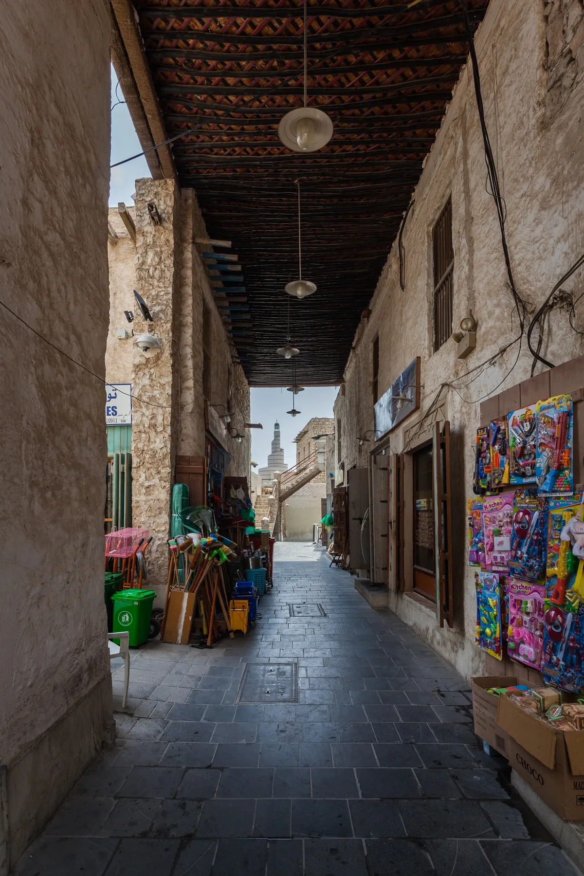 A narrow lane in Souq Waqif in Doha, mud-rendered walls under a wooden beamed roof with hanging lamps, stalls on either side and a spiral minaret visible at the far end
