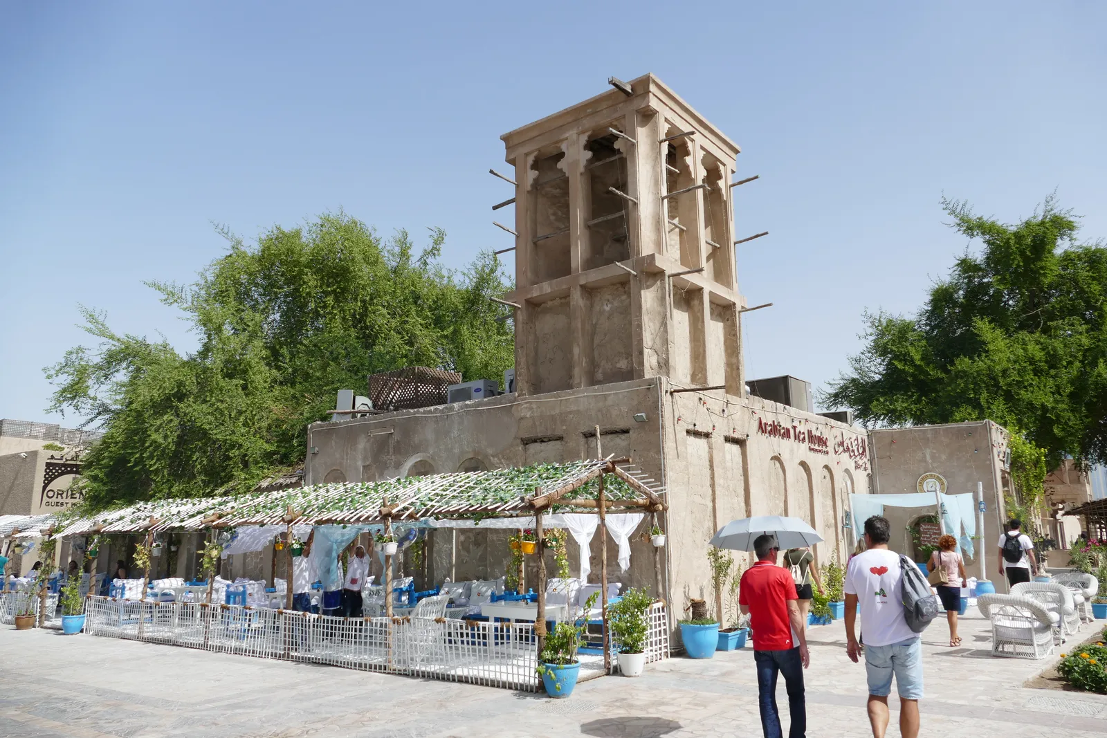 A square sand-coloured wind tower rising above a restored courtyard house in Dubai's Al Fahidi Historical Neighbourhood, with a shaded cafe terrace at its base