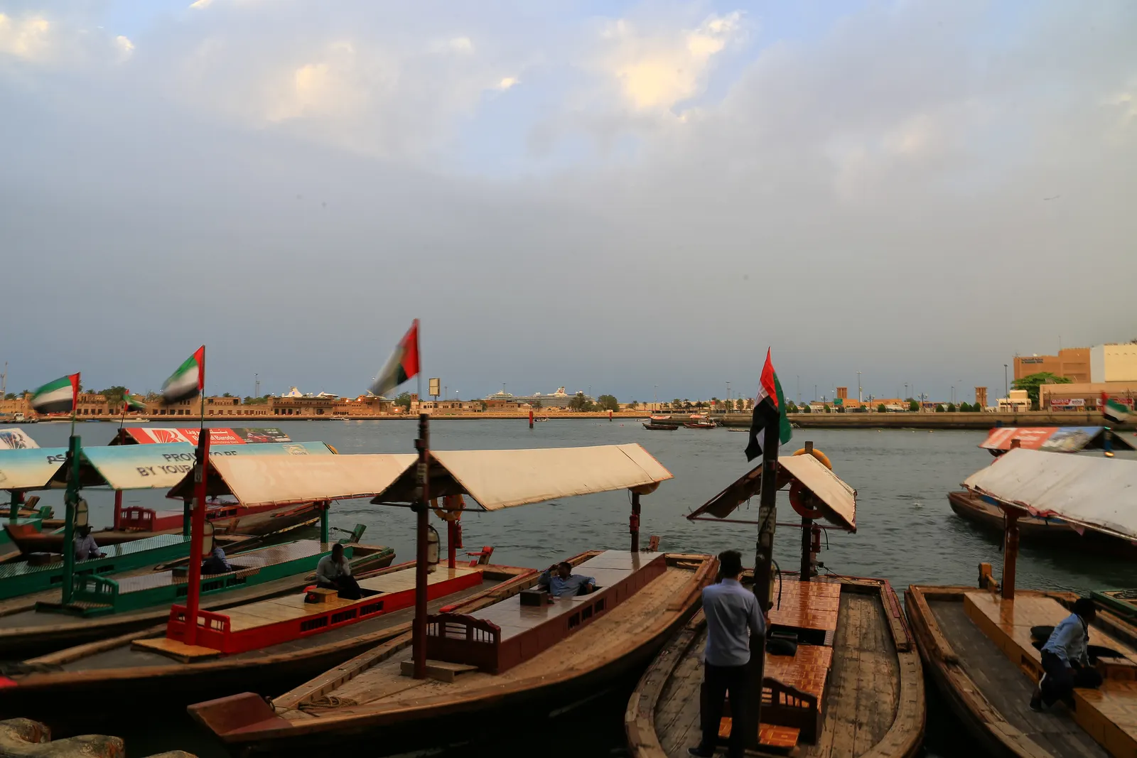 Wooden abra water taxis with canvas awnings and small UAE flags moored side by side at Bur Dubai Abra Station, with Dubai Creek and the Deira shore beyond