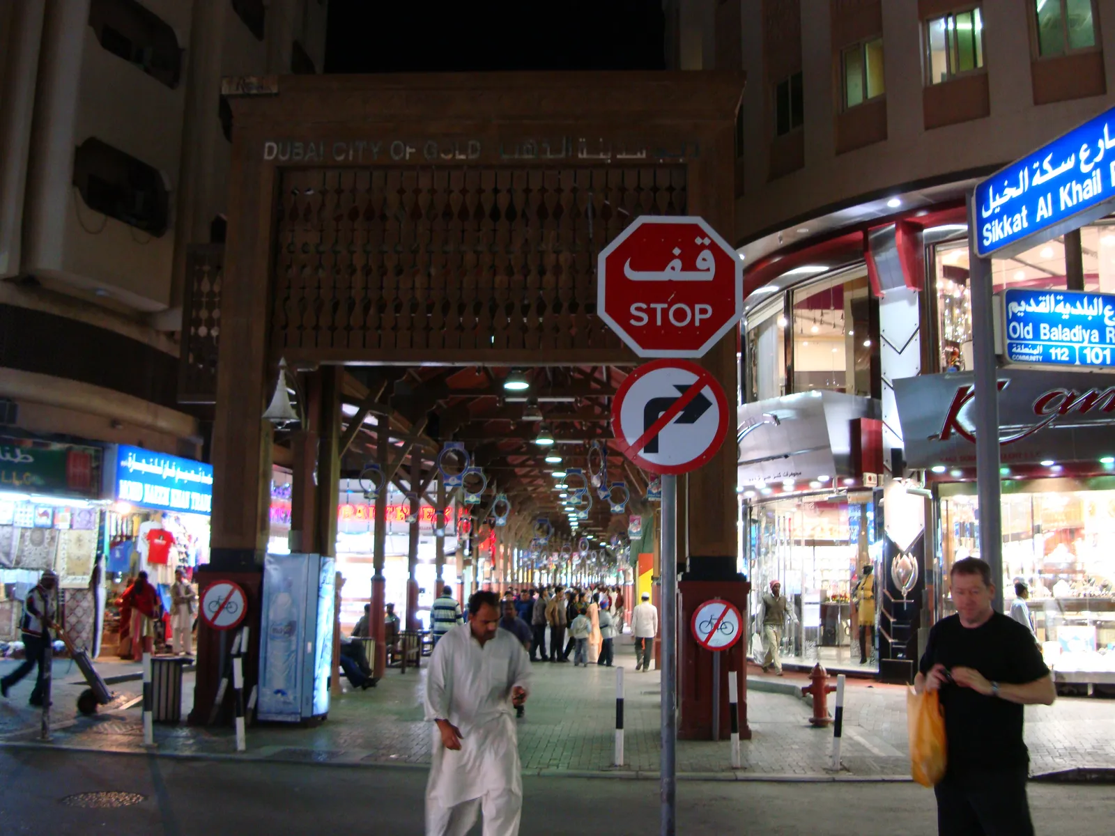The carved wooden entrance gate of the Dubai Gold Souk in Deira at night, its covered lane of jewellery shops lit up behind a street sign for Sikkat Al Khail
