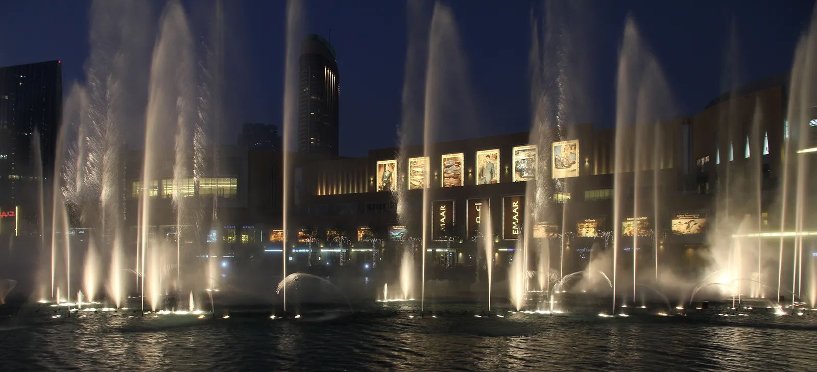Jets of the Dubai Fountain lit white against the dusk sky on Burj Lake, with the lit shopfronts of Downtown Dubai behind the water