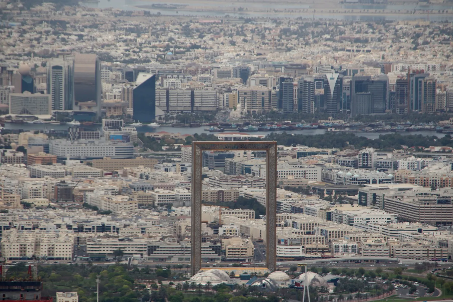 The Dubai Frame seen from high above, a tall rectangular gold-clad frame standing in Zabeel Park with the low-rise blocks of older Dubai and the creek behind it