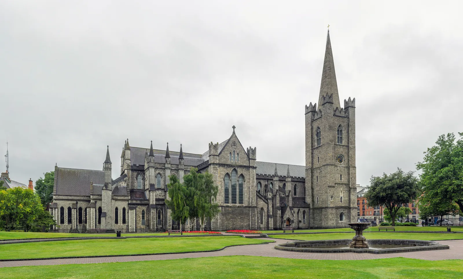 St Patrick's Cathedral in Dublin seen across its lawns, a long grey medieval church with a battlemented tower and stone spire at one end and a fountain on the grass in front
