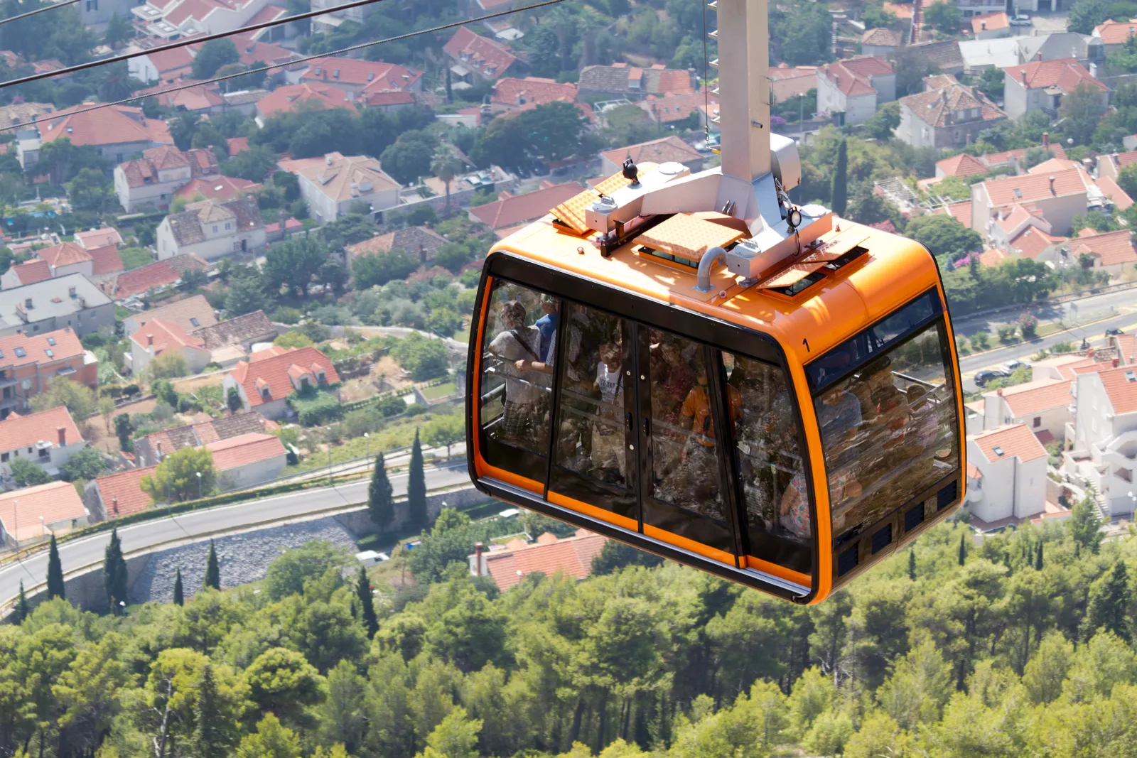 An orange cable car cabin climbing above Dubrovnik on its cable, with the tiled roofs and cypresses of the town spread out on the slope far below