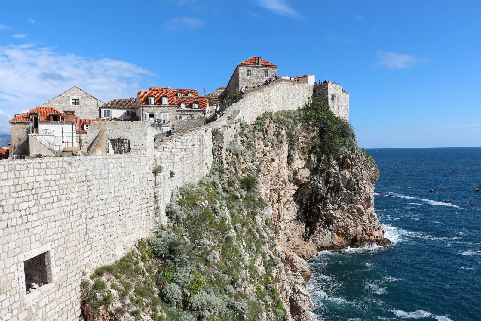 The seaward stretch of Dubrovnik's city walls running along the top of a limestone cliff above the Adriatic, with the roofs of the Old Town rising behind the rampart