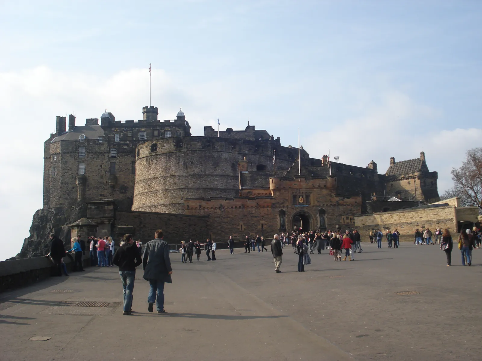 The esplanade at the top of Edinburgh's Royal Mile, with visitors crossing towards the castle gatehouse under the curved wall of the Half Moon Battery