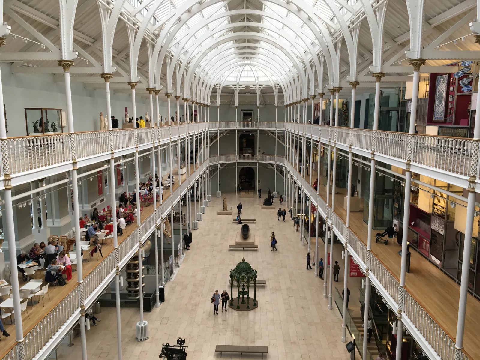 The Grand Gallery of the National Museum of Scotland, a long hall of white cast-iron columns and balconies under a glass barrel roof, with visitors on three levels