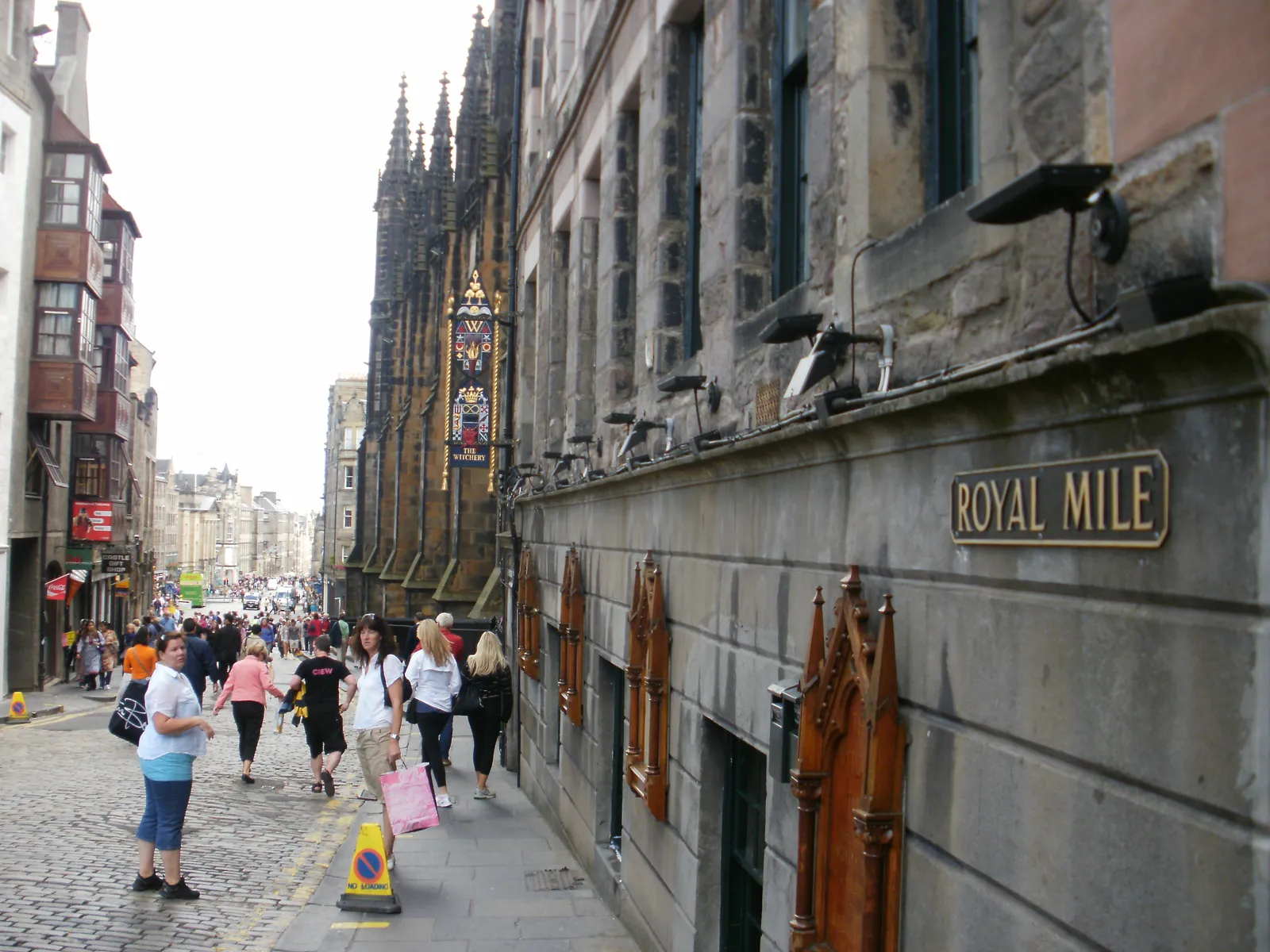 Pedestrians walking down the cobbled Royal Mile in Edinburgh's Old Town, past a street sign marking the route