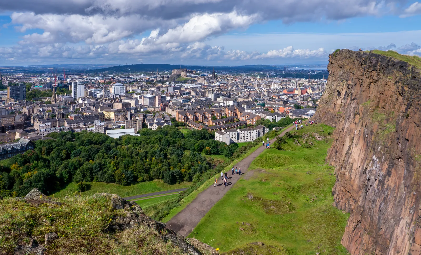 The dolerite cliffs of Salisbury Crags in Edinburgh's Holyrood Park, with a footpath running along the grass beneath them and the city spreading out to the horizon