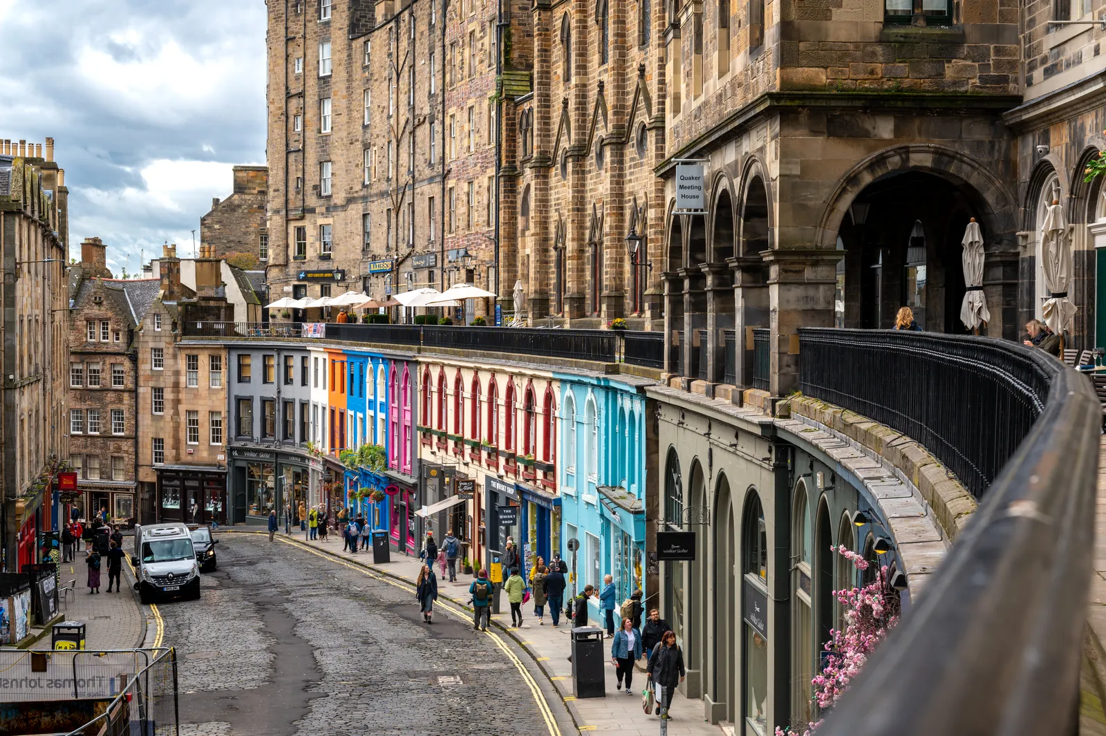 Victoria Street in Edinburgh curving downhill towards the Grassmarket, lined with brightly painted shopfronts, with the raised walkway of Victoria Terrace running above them