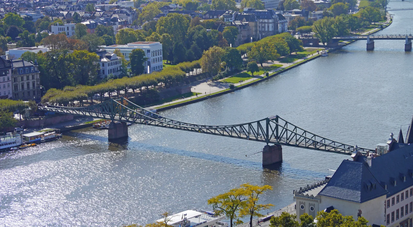 The Eiserner Steg, an iron lattice footbridge for pedestrians, crossing the river Main in Frankfurt between tree-lined embankments on both banks