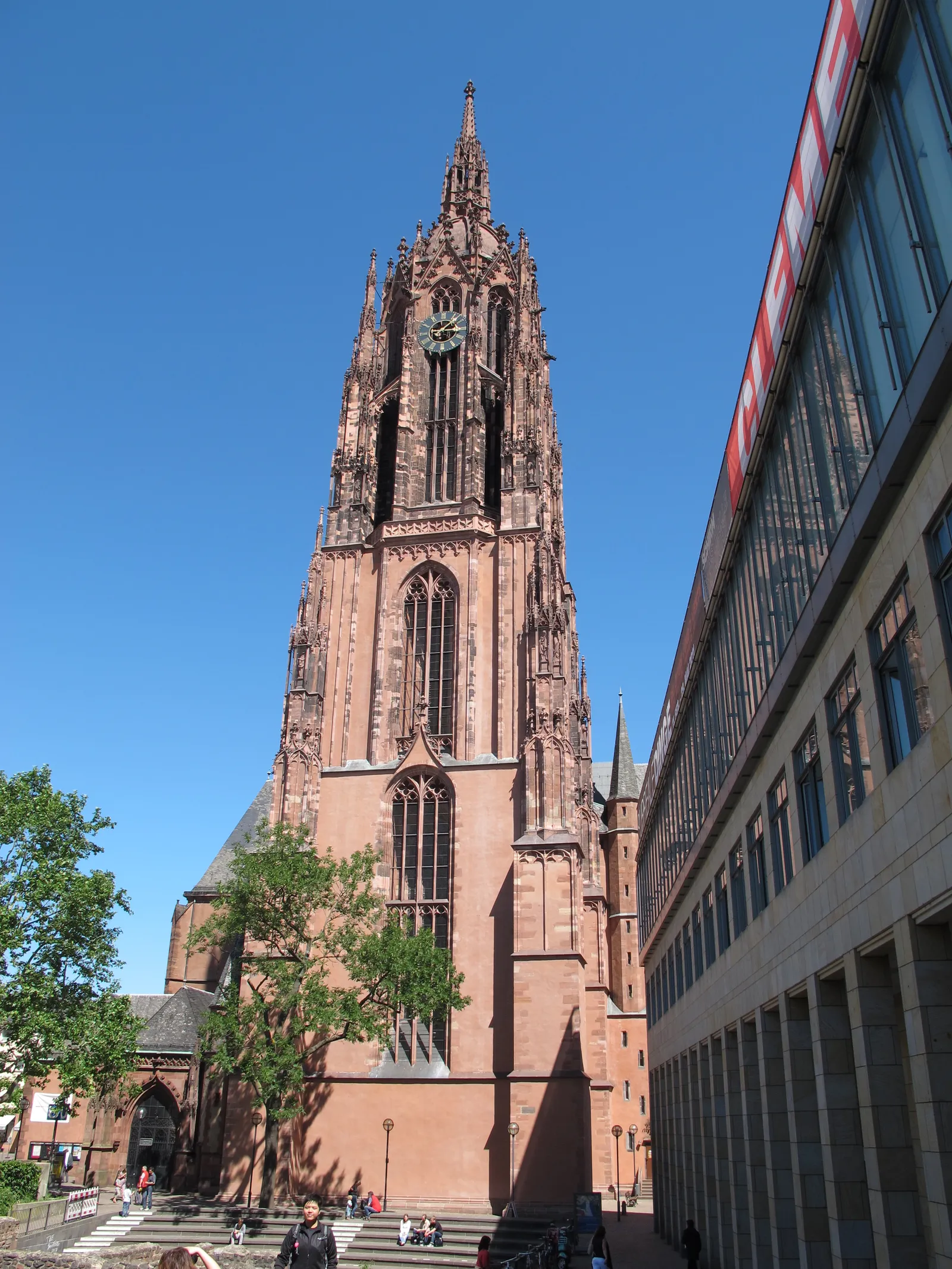 The red sandstone Gothic tower of Frankfurt Cathedral seen from the square below, its clock face and open stone lantern rising above the nave, with people sitting on the steps at its foot