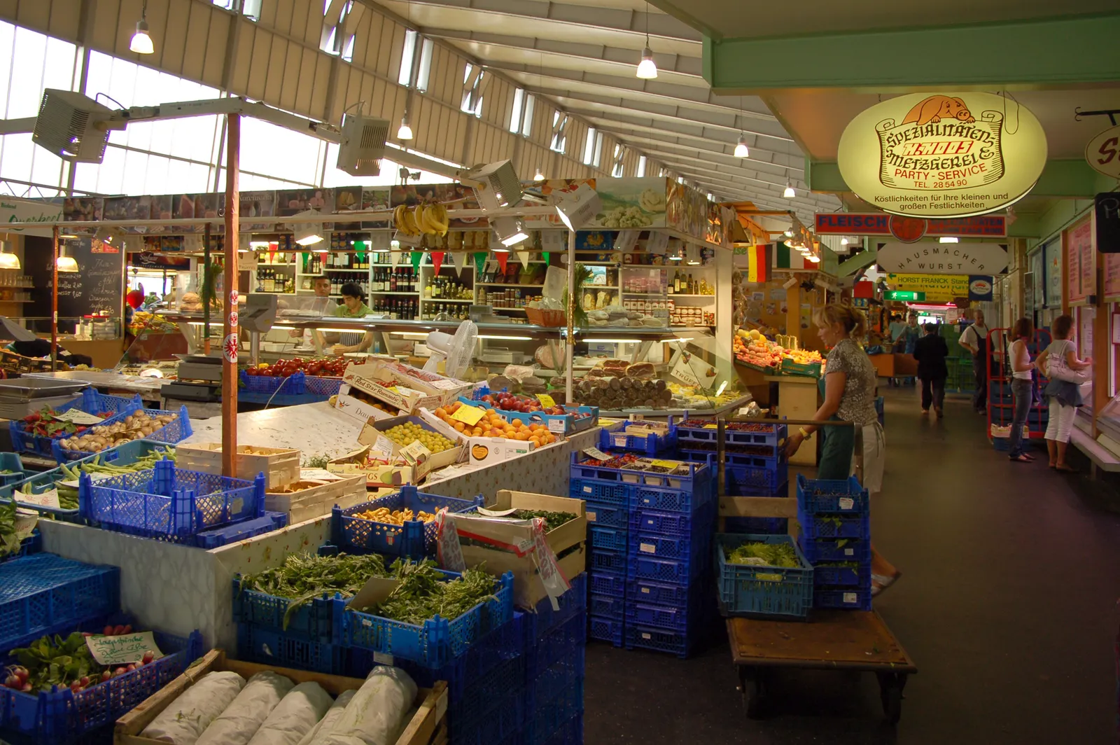 Inside Frankfurt's Kleinmarkthalle, stacked crates of vegetables in the foreground and food stalls running the length of the hall under a pitched roof with high windows