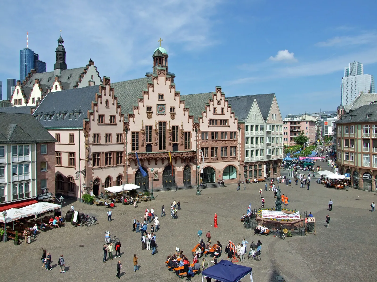 The Römerberg square in Frankfurt seen from above, the three stepped gables of the Römer city hall filling the far side, gabled and half-timbered houses to the right, and people crossing the paved square between them