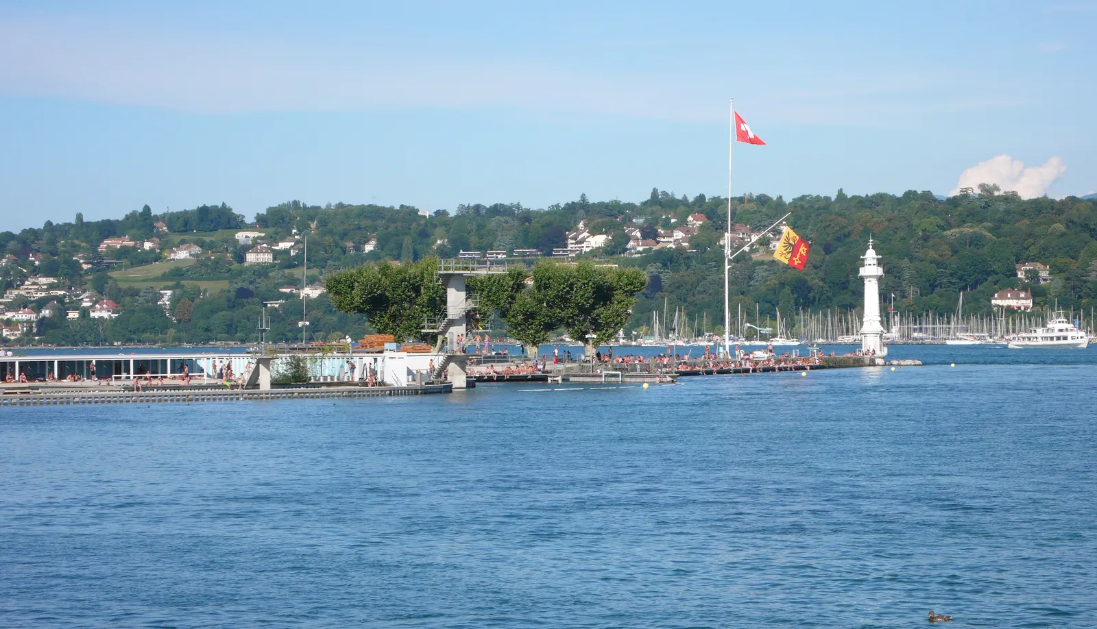 The Bains des Pâquis pier reaching out into Lake Geneva, swimmers along its edge and a small stone lighthouse at the far end under a Swiss flag