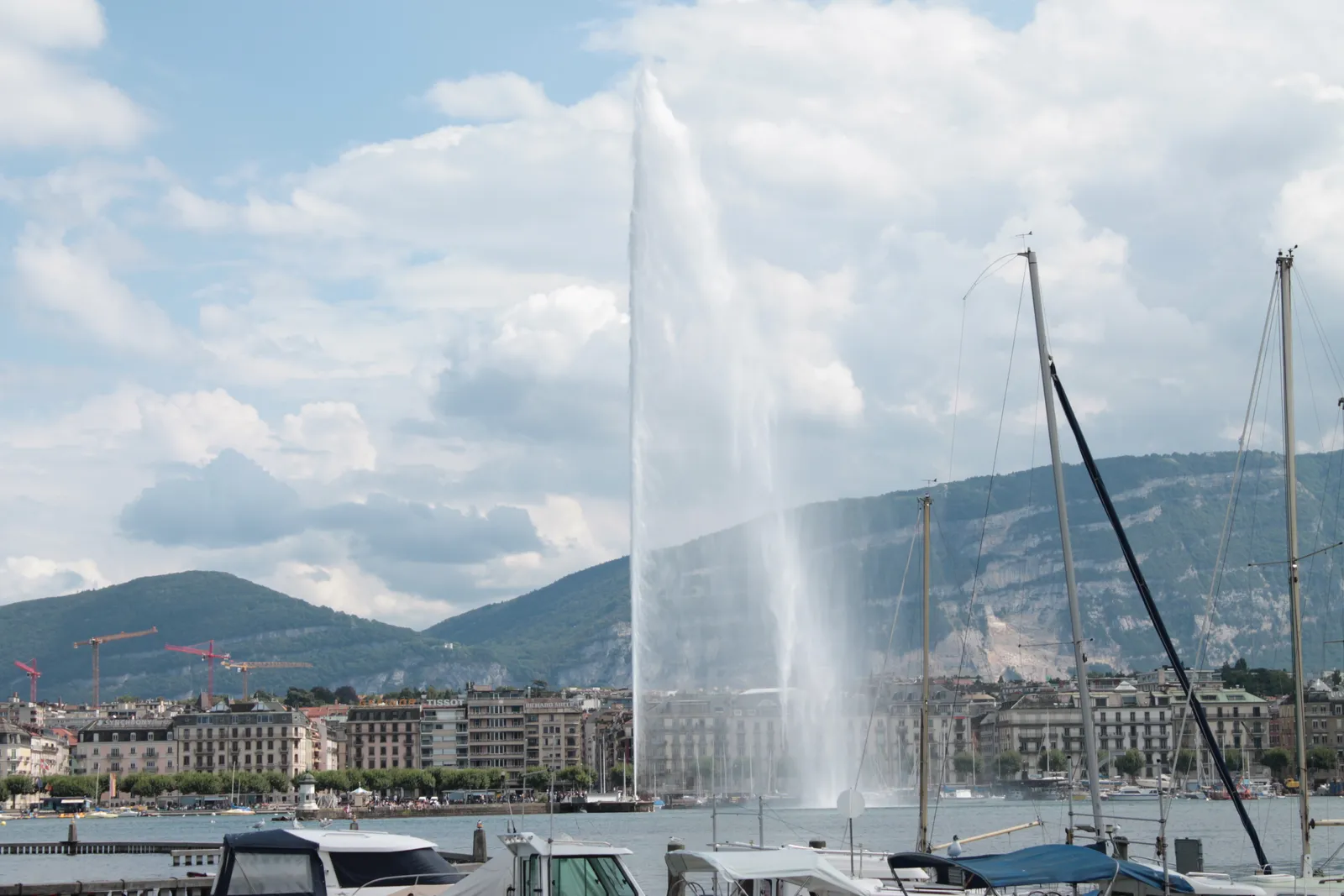 The Jet d'Eau throwing a tall white column of water above Geneva's harbour, moored yachts and their masts in the foreground and wooded hills along the far shore