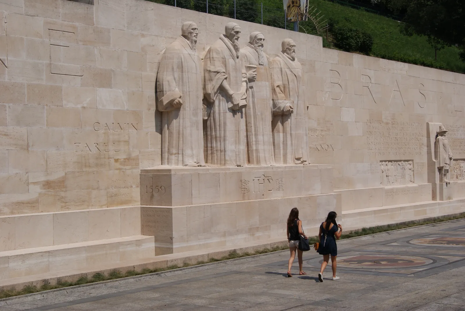 The Reformation Wall in Geneva, four tall stone figures of reformers carved in relief along a pale limestone wall with their names cut into the blocks beside them