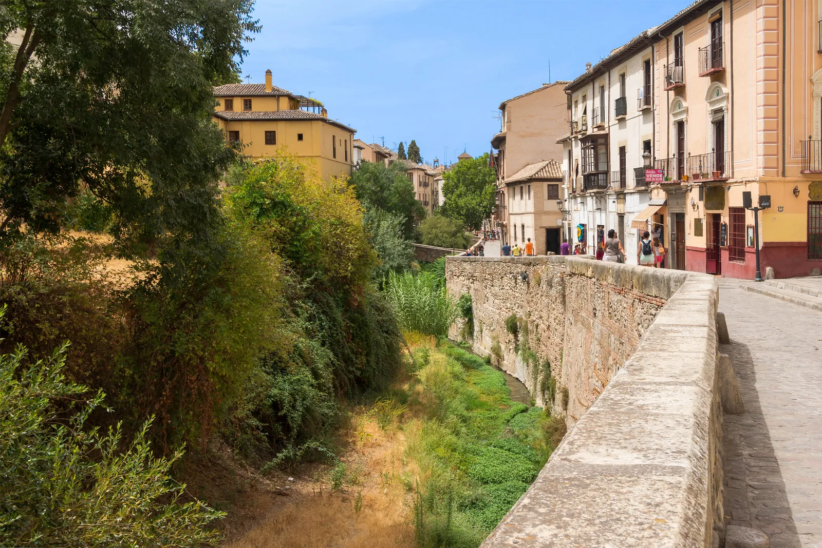 Carrera del Darro in Granada, a narrow paved lane with a low stone wall above the overgrown Darro riverbed and tall painted houses with balconies on the other side