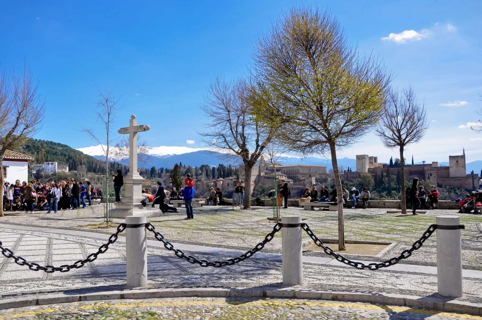 The Mirador de San Nicolás in Granada, a paved terrace with a stone cross and people sitting along the wall, the Alhambra on its hill beyond and snow on the Sierra Nevada behind