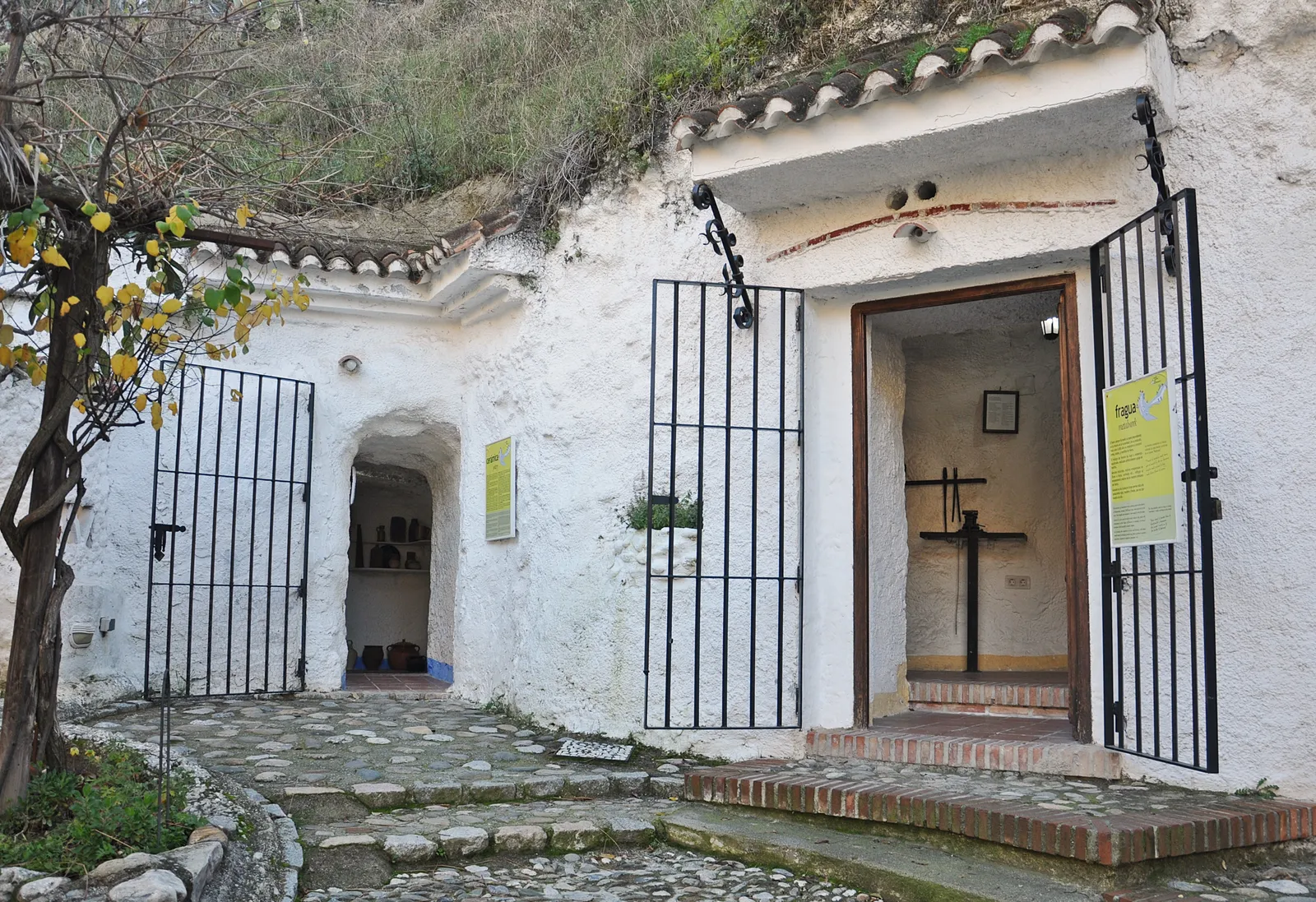 Whitewashed cave-house fronts at the Sacromonte cave museum in Granada, iron gates across the doorways and the bare hillside rising directly above the roofs