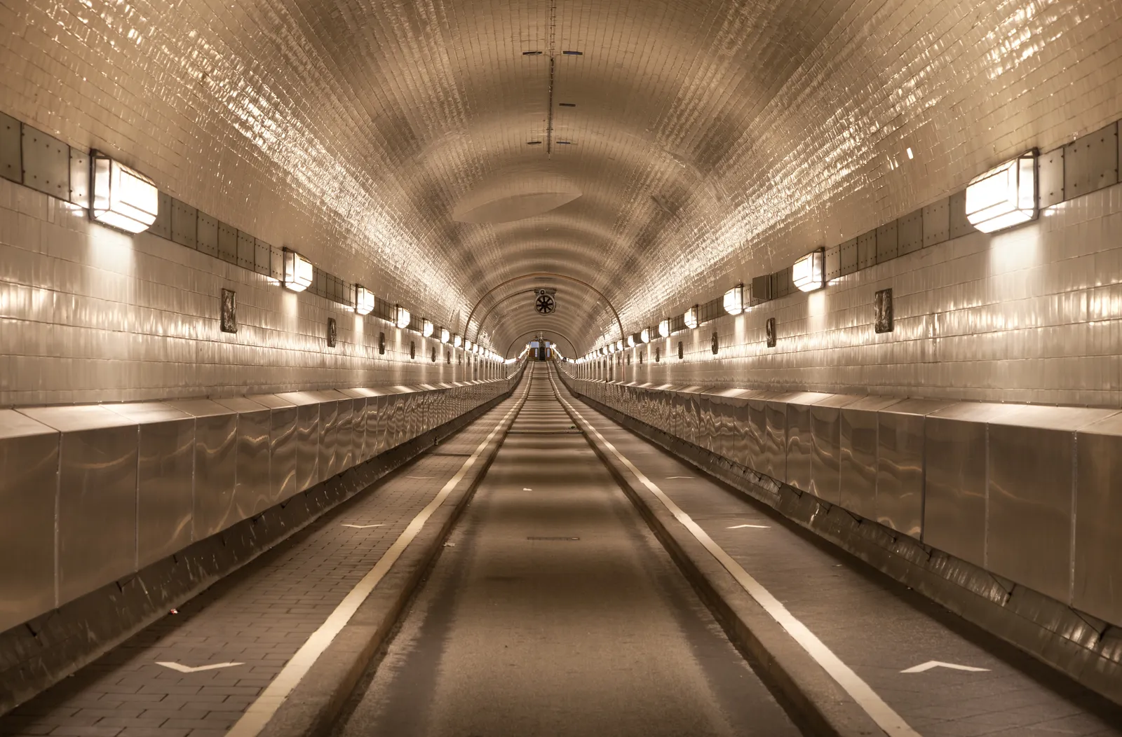 The empty white-tiled tube of Hamburg's Old Elbe Tunnel running away into the distance, lit by wall lamps along both sides