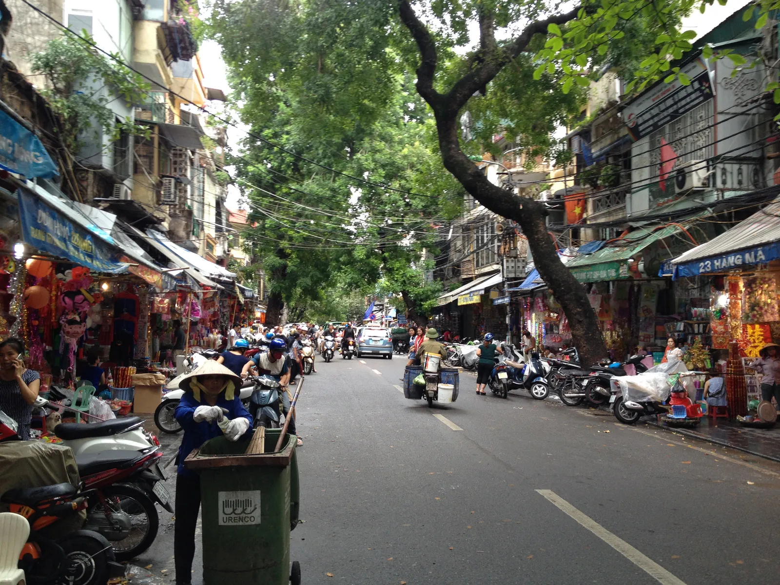 A narrow street in Hanoi's Old Quarter lined with shophouses, their goods stacked out onto the pavement under awnings, with parked motorbikes along both kerbs and trees overhanging the road