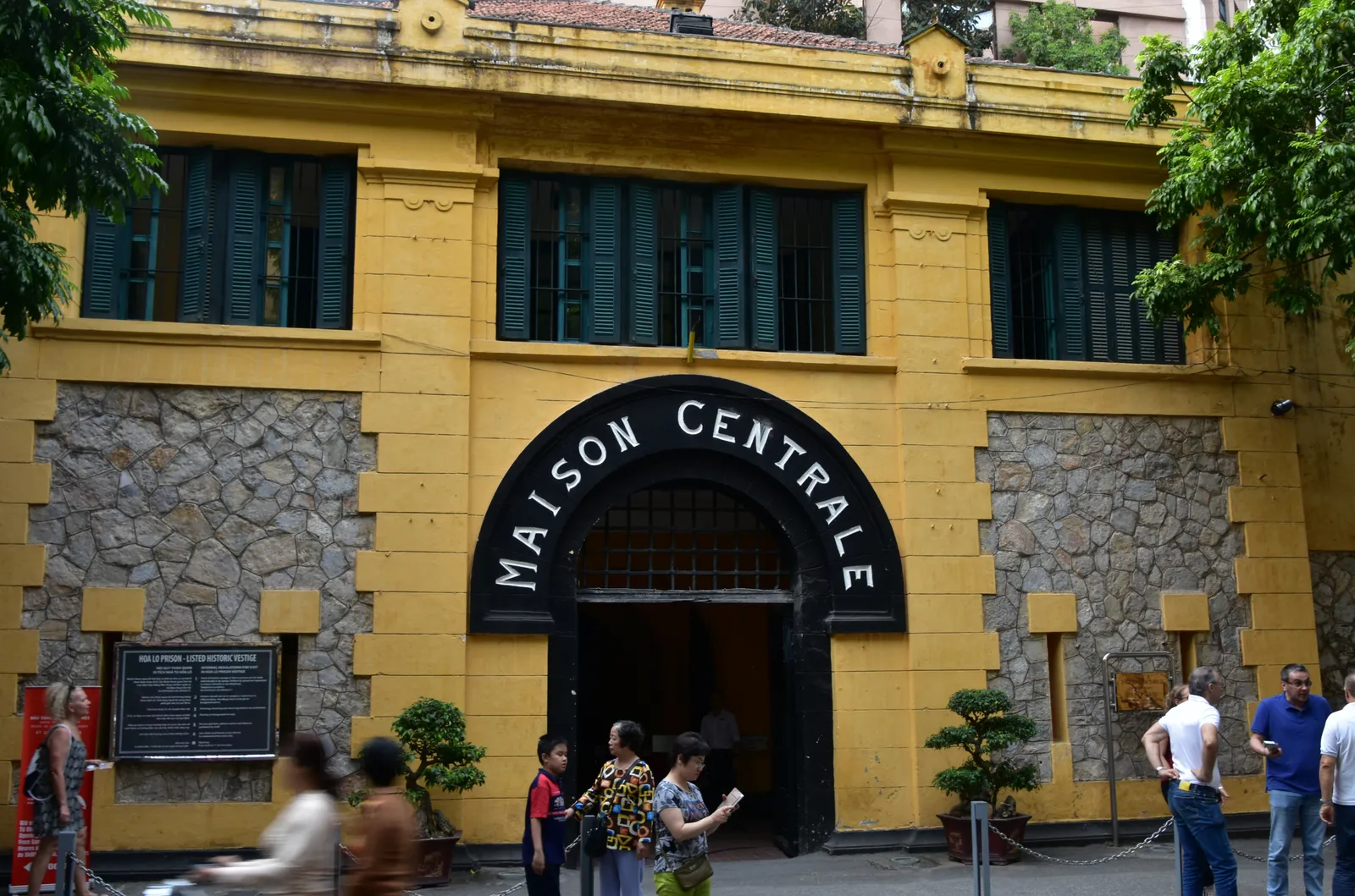 The yellow gatehouse of Hoa Lo Prison in Hanoi with the words Maison Centrale arched in black lettering above the entrance and visitors walking in off the street