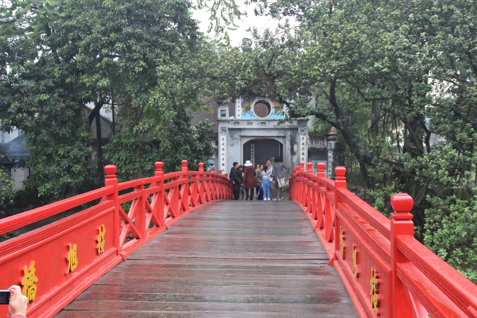 The red wooden Huc Bridge in Hanoi curving across the water to the stone gateway of Ngoc Son Temple, with visitors walking over it under overhanging trees