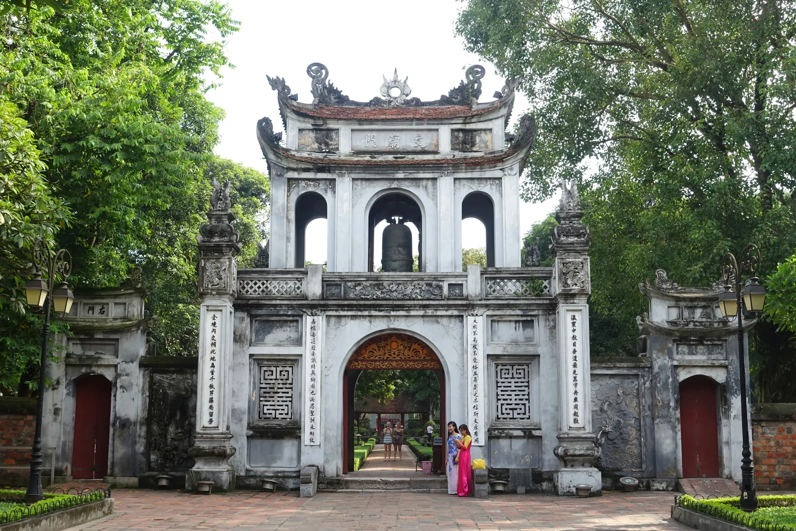 The grey stone entrance gate of the Temple of Literature in Hanoi, two storeys with a small bell chamber above the central arch, with two women in pink and blue dresses standing in the archway