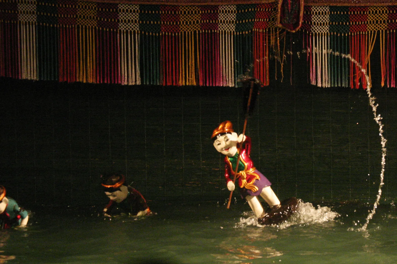 A lacquered water puppet of a farmer poling across the flooded stage at the Thang Long theatre in Hanoi during the rice-planting scene, with a second puppet in the water behind it