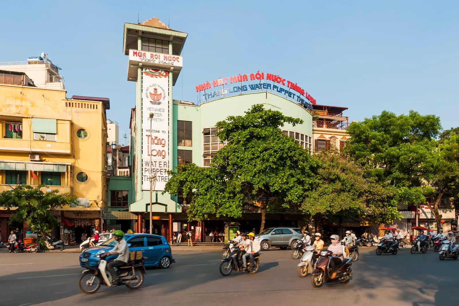 The Thang Long Water Puppet Theatre in Hanoi, a green building with a tall red and yellow sign tower, with motorbikes and cars crossing the junction in front of it