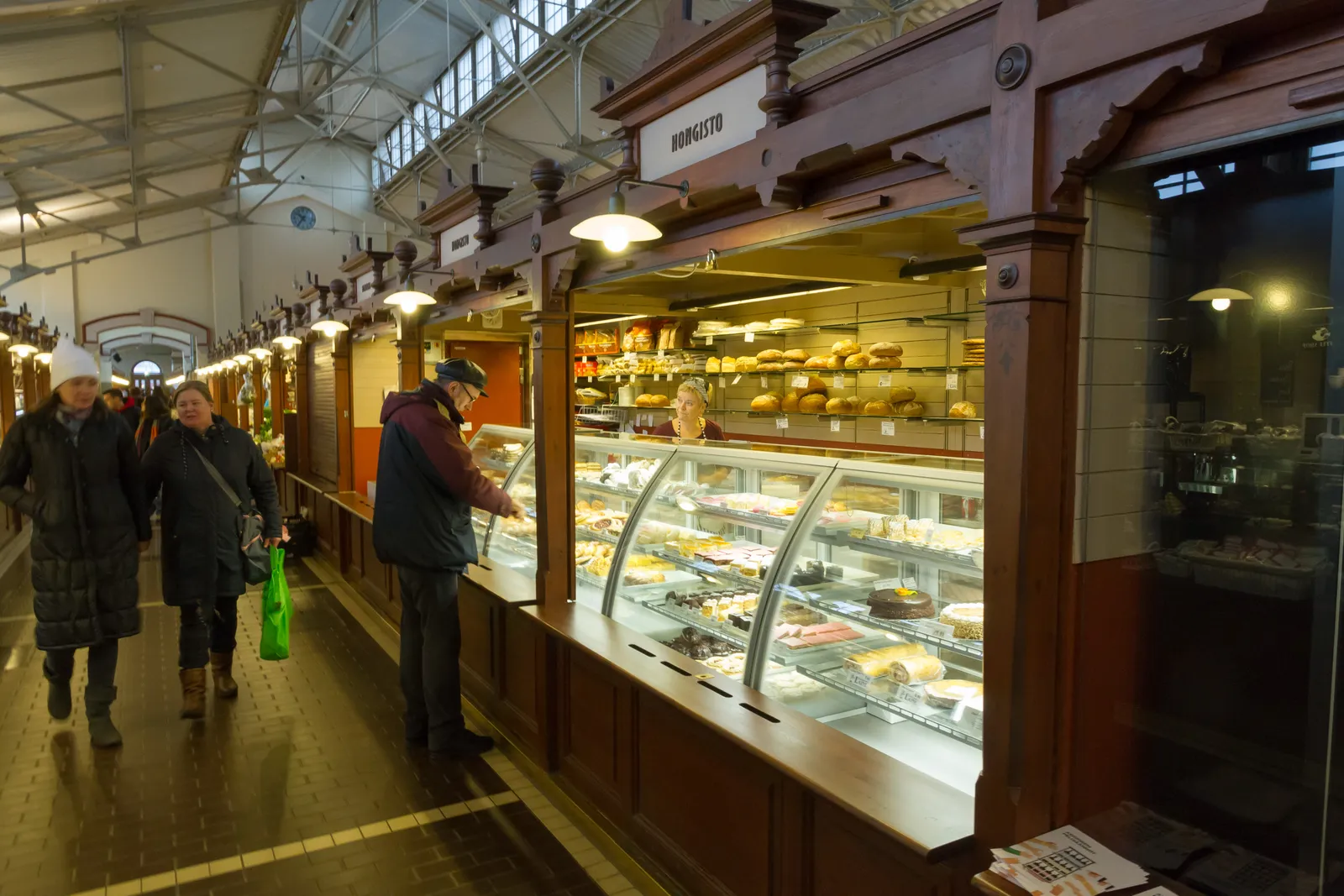The aisle inside Helsinki's Old Market Hall, dark timber stall fronts with glass counters and hanging lamps down one side under an arched iron and glass roof, with shoppers walking between them