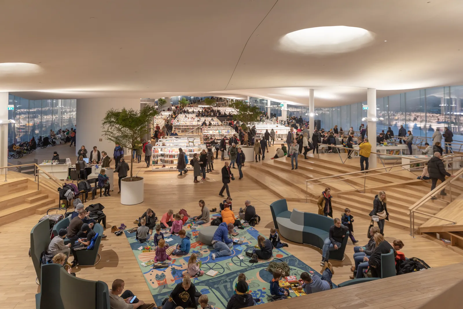 The top floor of Oodi library in Helsinki, a wide timber-floored room under a curving white ceiling with round skylights, low bookshelves and armchairs spread across it and a glass wall along one side