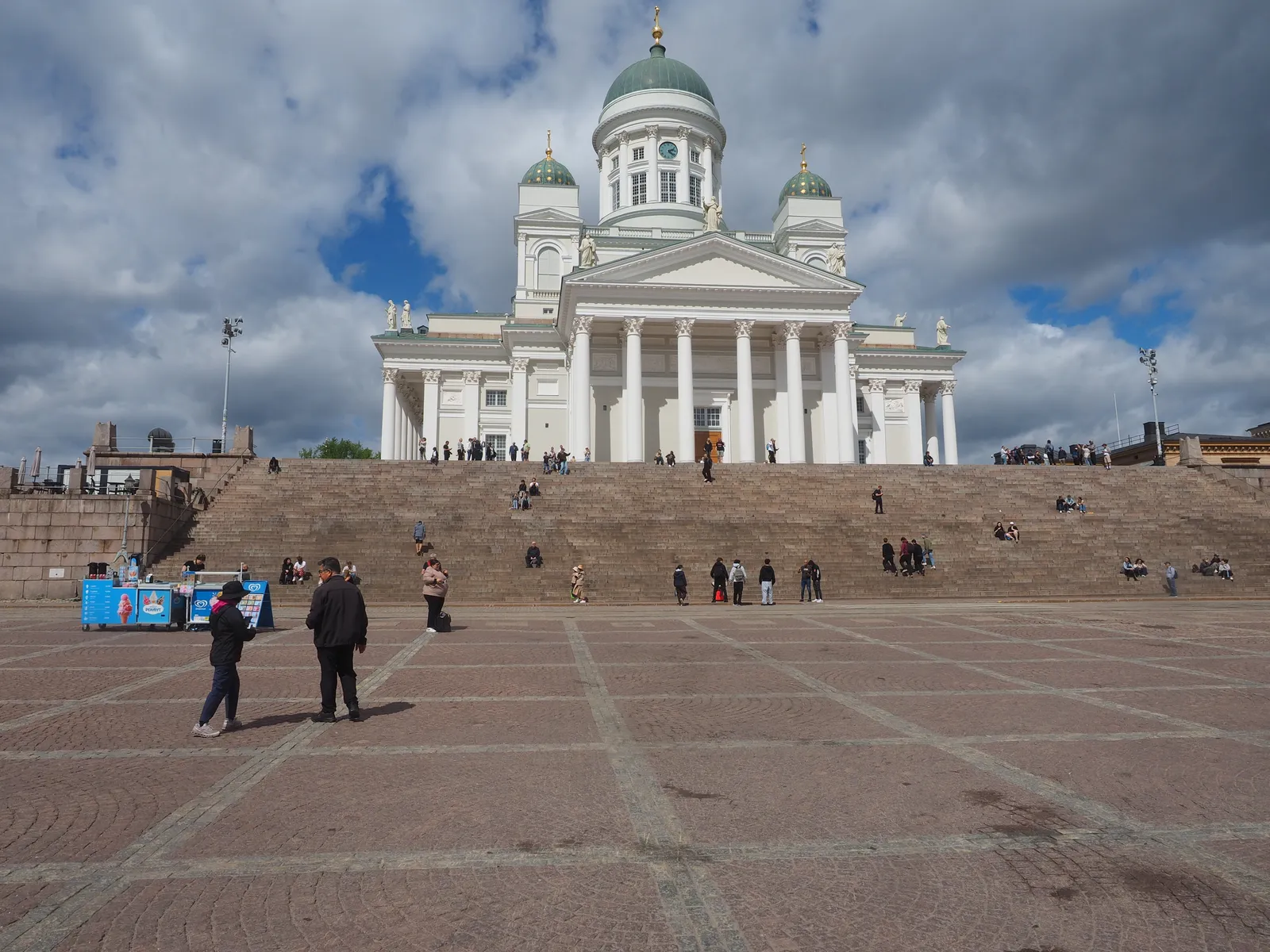 Helsinki Cathedral at the top of its wide stone staircase on Senate Square, a white neoclassical church with a green central dome and four smaller ones, people sitting on the steps and walking across the cobbled square below