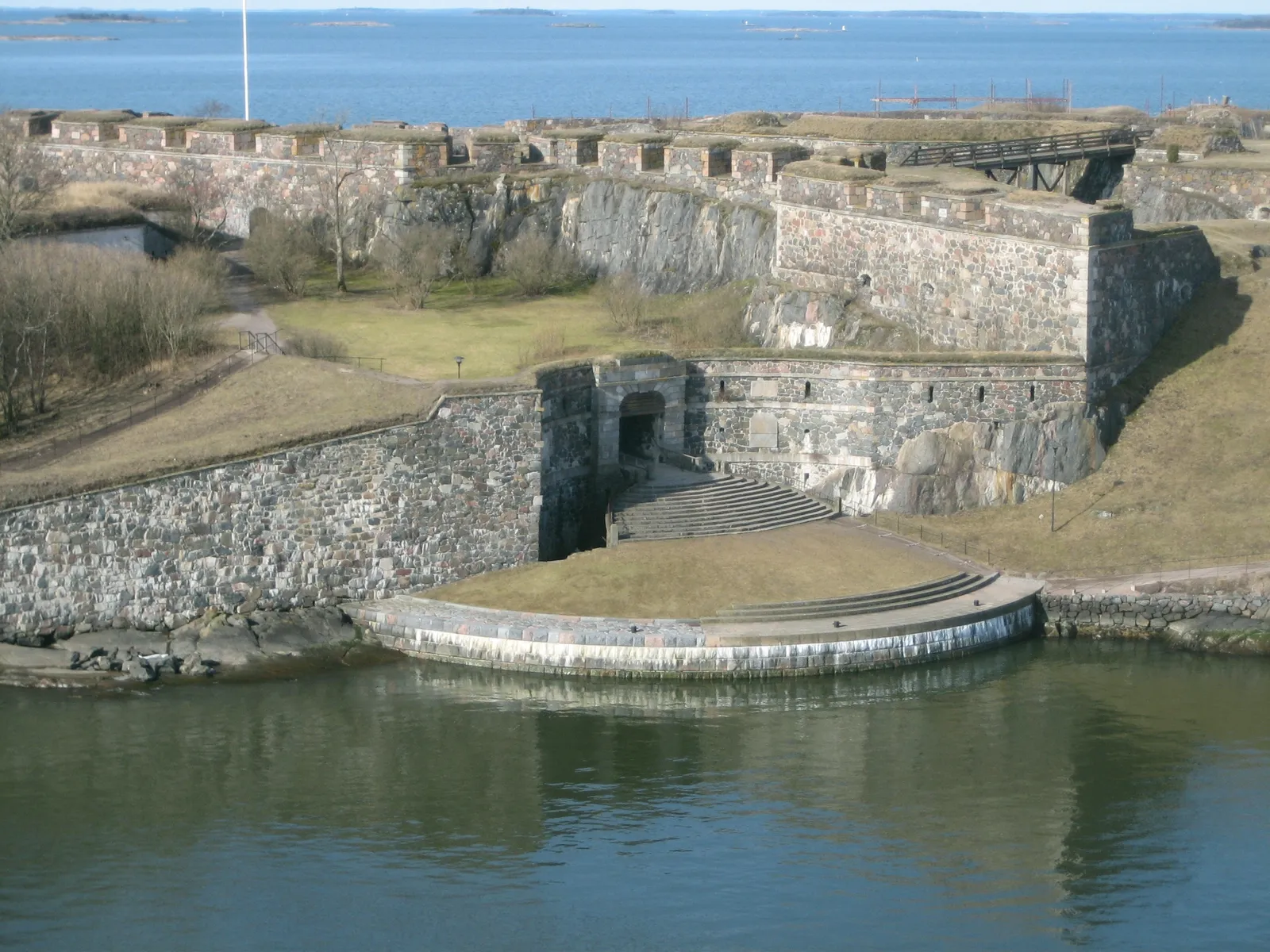 The King's Gate at Suomenlinna seen from the water, a stone archway set into a grass-topped granite rampart with a broad flight of steps running down from it to a curved quay at the sea's edge, open water and low islands behind