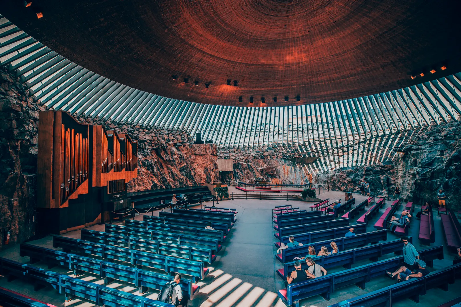 The inside of Temppeliaukio Church in Helsinki, rough grey rock walls rising to a shallow copper dome ringed by a band of skylights, with an organ against the left wall and rows of pews on the stone floor