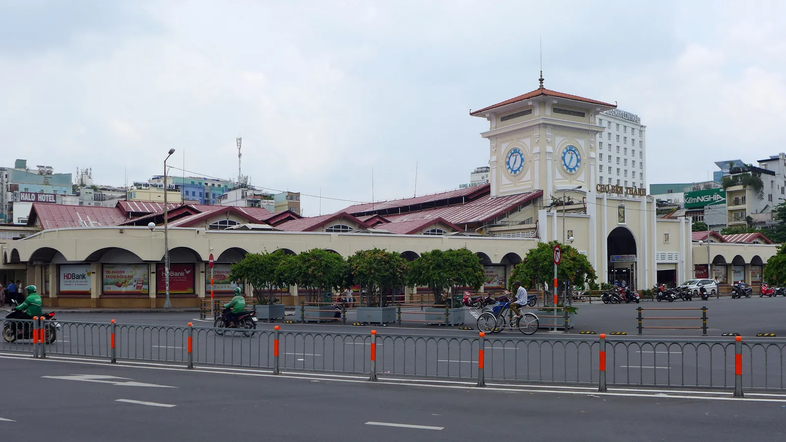 The south gate of Ben Thanh Market in Ho Chi Minh City with its square clock tower, the long tiled market hall stretching away behind it and motorbikes crossing the roundabout in front