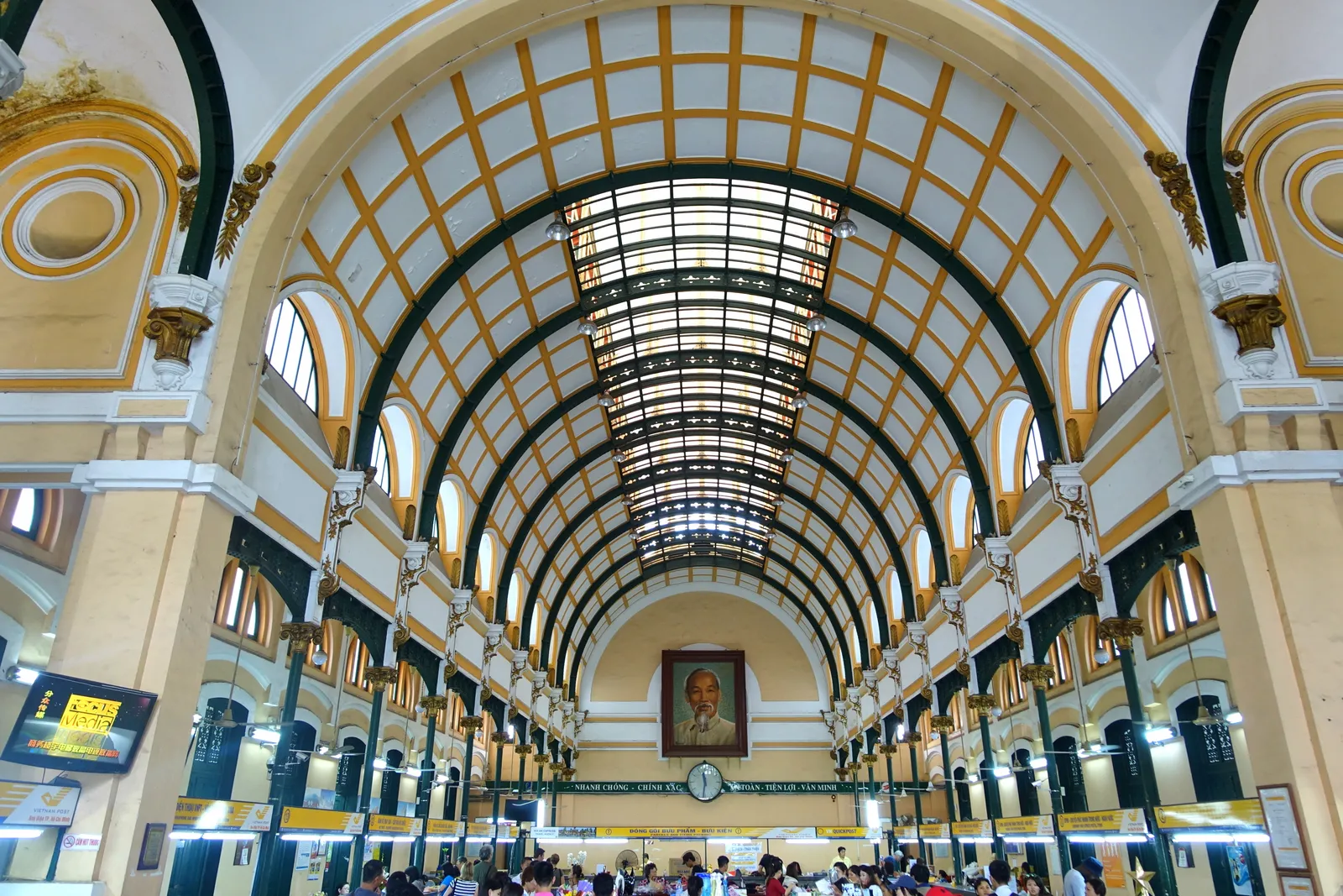 The main hall of Saigon Central Post Office under its glazed barrel vault, green iron arches running the length of the ceiling and a portrait of Ho Chi Minh on the end wall above the counters
