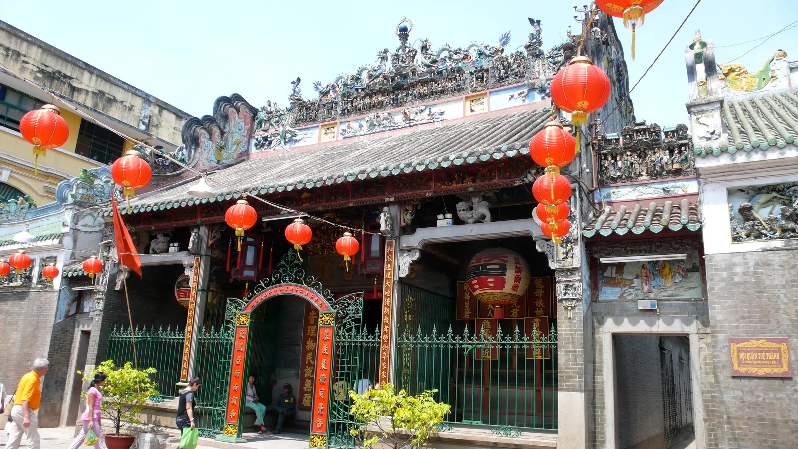 The entrance of Thien Hau Temple in Cho Lon, red paper lanterns strung across the forecourt and the roof ridge crowded with small glazed ceramic figures above the green iron gates